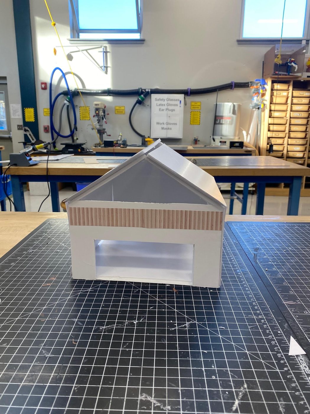A white foam board tiny house model with a gable roof, a printed wood siding band and a large clear window